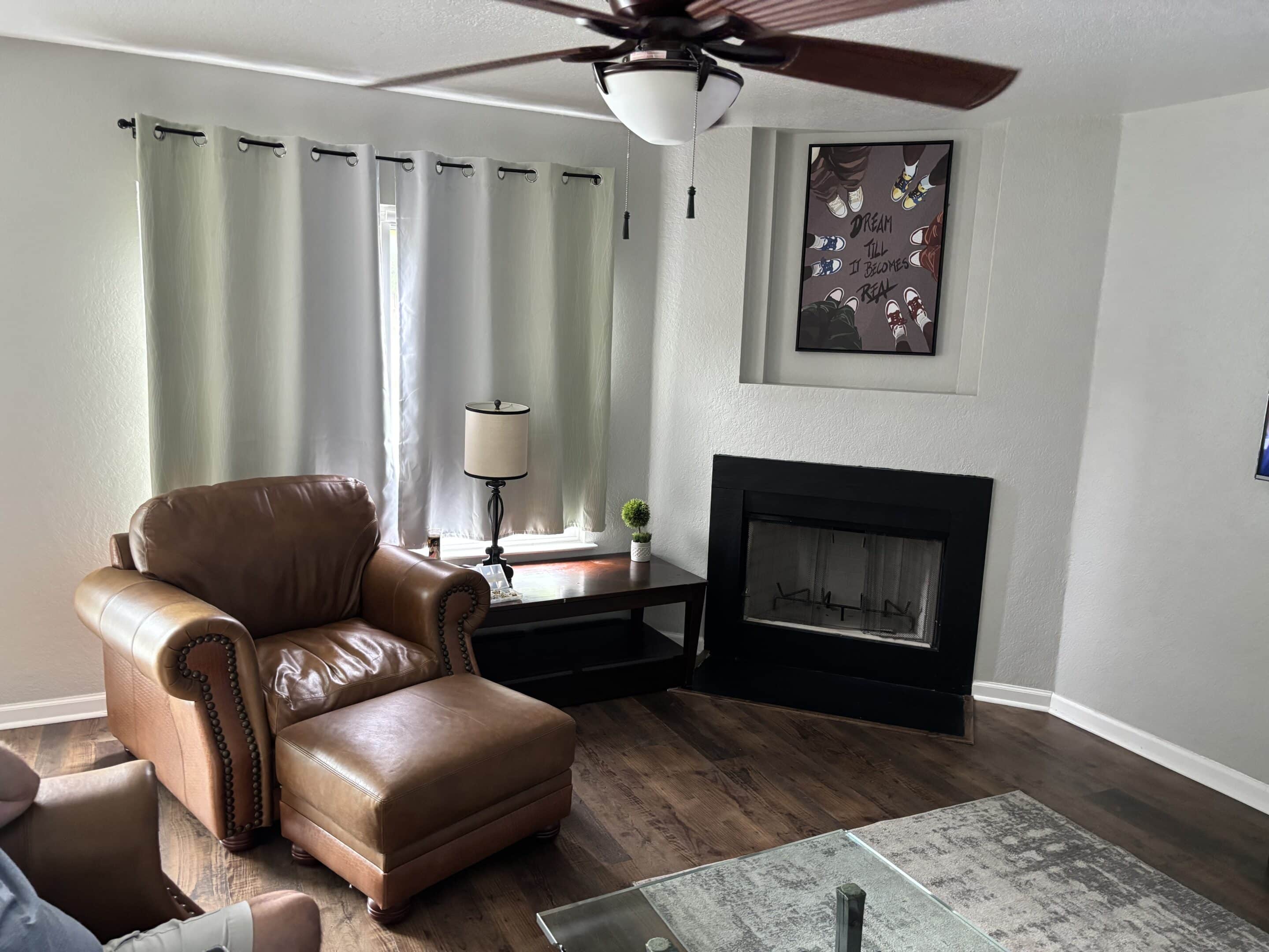 Cozy living room corner of a sober-living home at 7263 Dwarf Palm—featuring a leather armchair with ottoman, side table with lamp, fireplace with framed artwork above, ceiling fan, and hardwood floors, all bathed in natural light.
