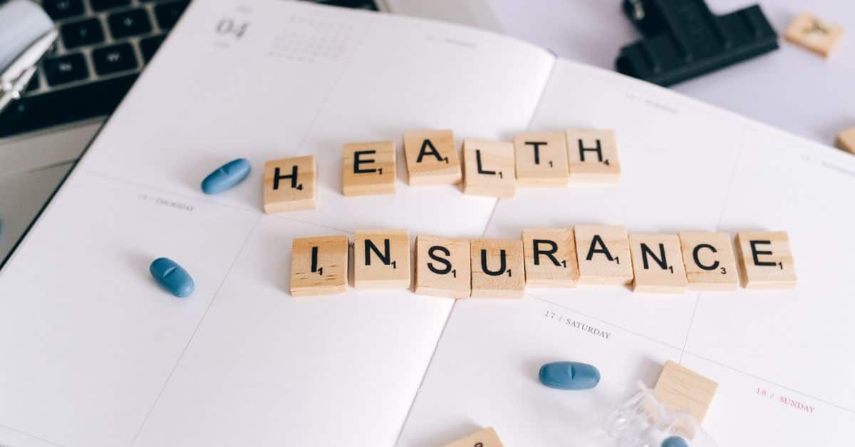 Scrabble tiles spelling 'Health Insurance' on planner with pills and laptop, symbolizing healthcare planning.