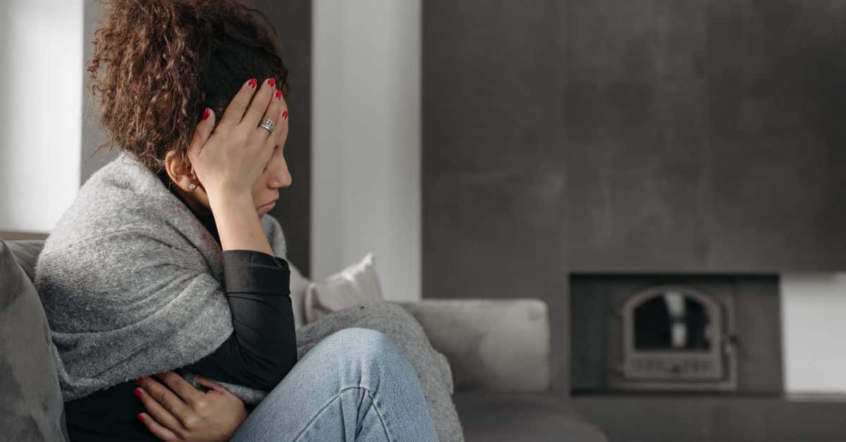 A woman sits on a couch indoors, wrapped in a blanket, holding her head in discomfort.