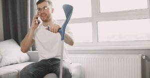 Adult man sitting on couch using crutch, talking on phone in a well-lit room.