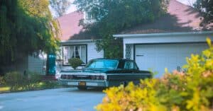 A classic black Chevrolet Impala parked in a suburban driveway surrounded by greenery.