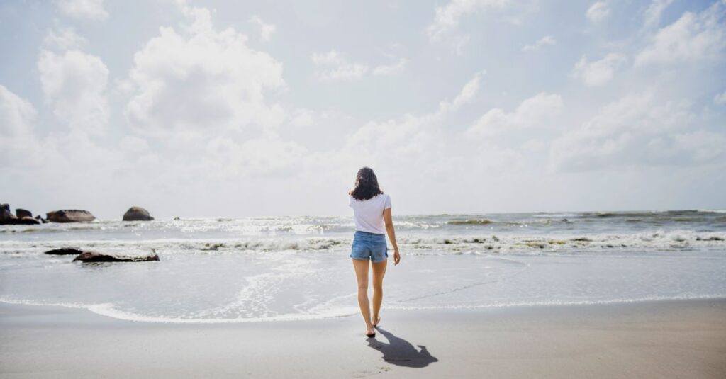 A woman enjoys a peaceful walk on a sandy beach with gentle waves and cloudy skies.