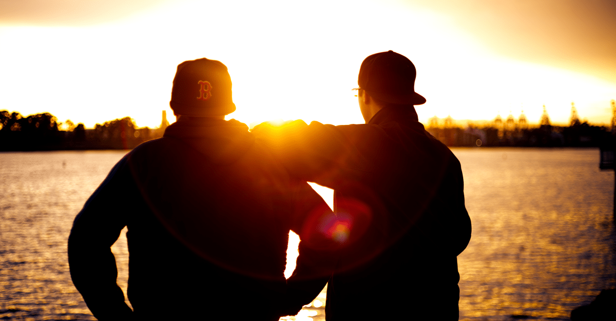 Two friends in baseball caps watch a sunset over the water, creating a serene silhouette.