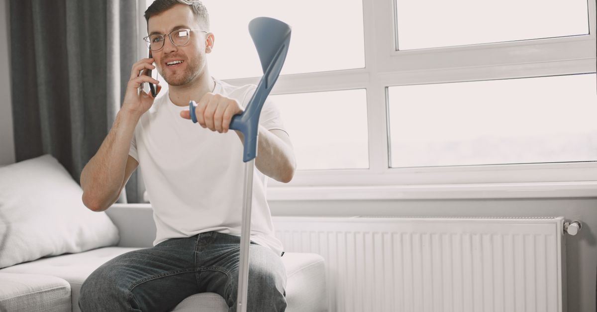 Adult man sitting on couch using crutch, talking on phone in a well-lit room.