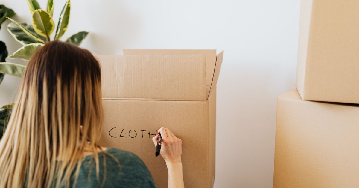 Back view anonymous female in casual outfit writing on cardboard container word cloth while packing belongings and preparing to move