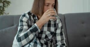 A man with long hair wears a checkered shirt, sitting on a couch, drinking water.