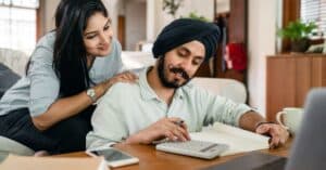 Concentrated ethnic male accountant using calculator with gadgets and books and notebook for remote work while ethnic wife touching shoulder of husband watching for numbers behind back sitting on sofa