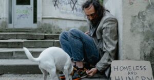 A homeless man sits on a street with his dog beside a sign reading 'Homeless and Hungry'.
