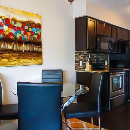 Glass-top dining table and chairs beside a modern kitchen with dark cabinets and stainless appliances.